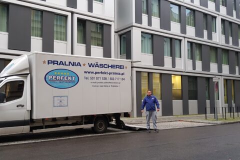 Samochód dostawczy Pralni Perfekt z pracownikiem przed budynkiem hotelowym, gotowy do dostawy